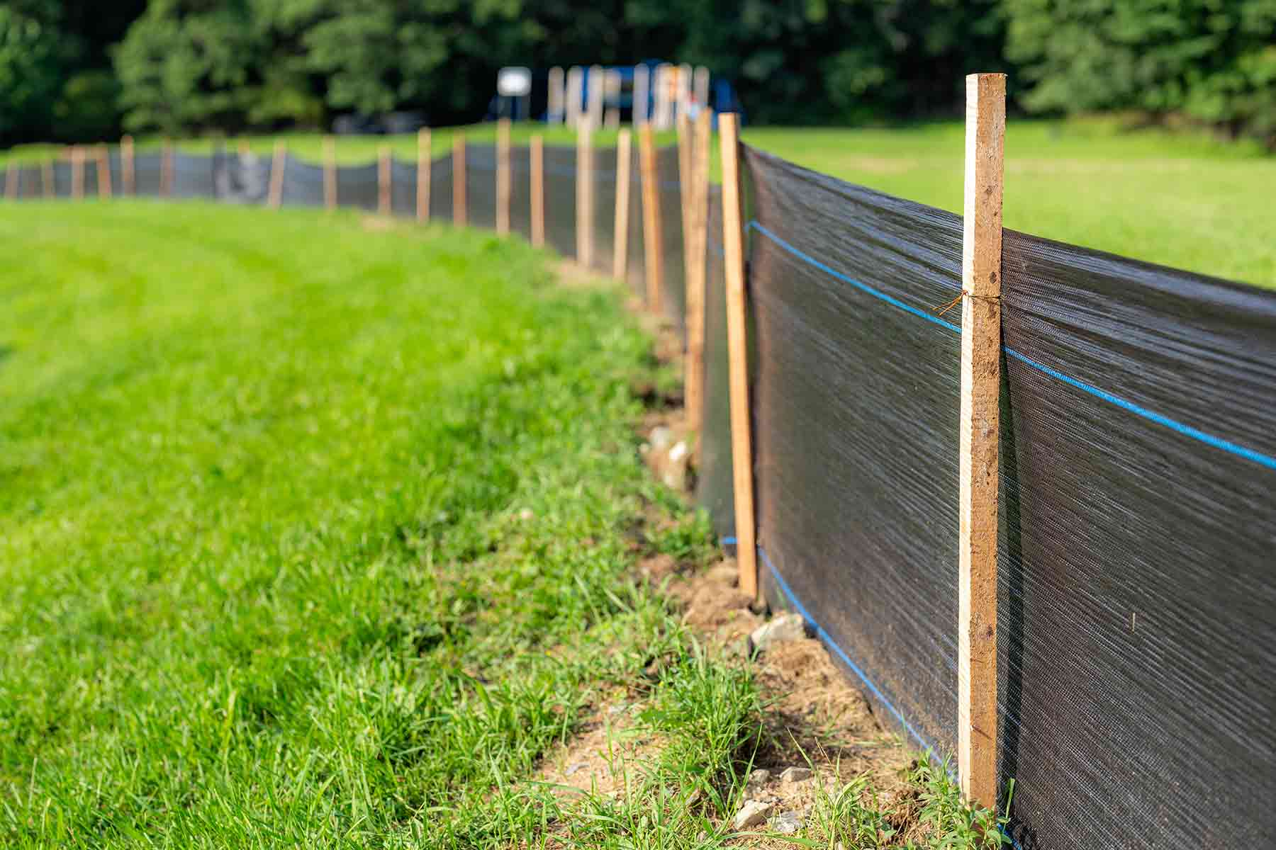 Black silt fence fabric trenched and stapled to wooden stakes along the edge of a green field